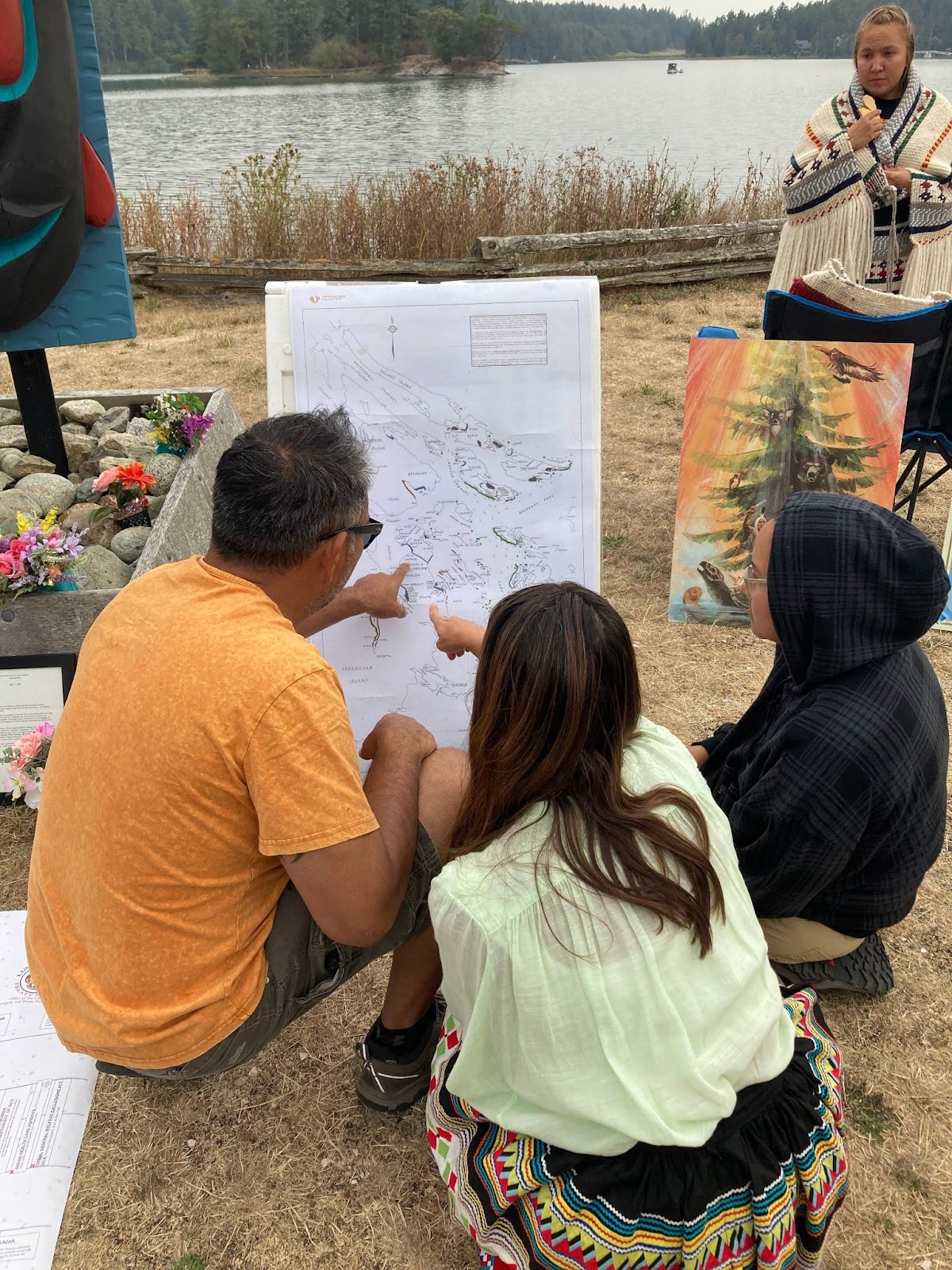 Three people sit in front of a large map printed on white paper, while two of them point at different areas
    on the map. In the background a person wearing a Coast Salish woven shawl looks on while water and trees across
    the water can be seen in the distance.