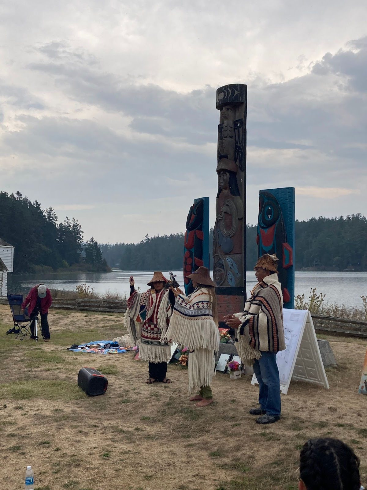 Three people wearing Coast Salish woven clothing and cedar hats stand in front of 3 story poles with their
    hands raised in front of their bodies. One of them holds a rattle in each hand.
    In the background water and trees can be seen.