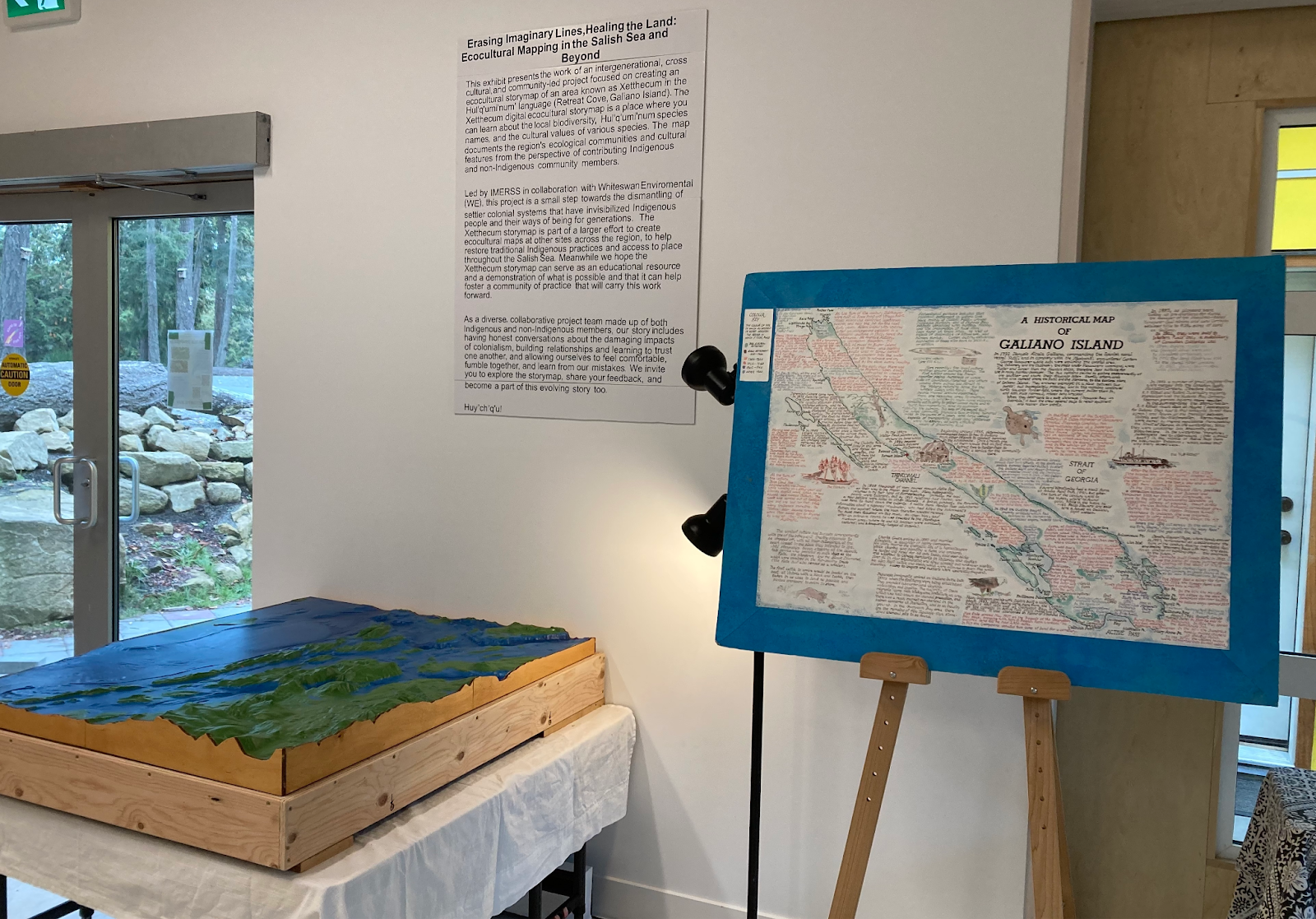 Beside an entrance door, a 3D map sits on a table in front of text mounted on the wall. Another map of Galiano
    Island with text and illustrations can be seen mounted on an easel.