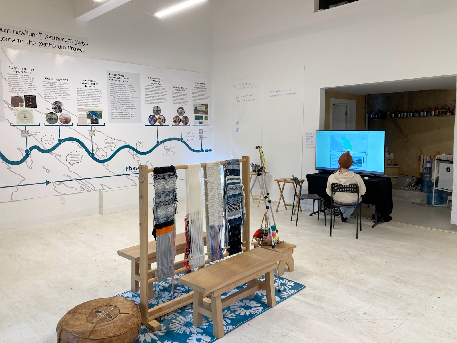 A view inside the gallery showing the timeline banner on the wall to the left, the Coast Salish loom in the
    centre of the room, someone sitting in front of the computer screen on which can bee seen a map, and large pieces of
    paper mounted on the wall beside the computer screen with sticky notes stuck to them.