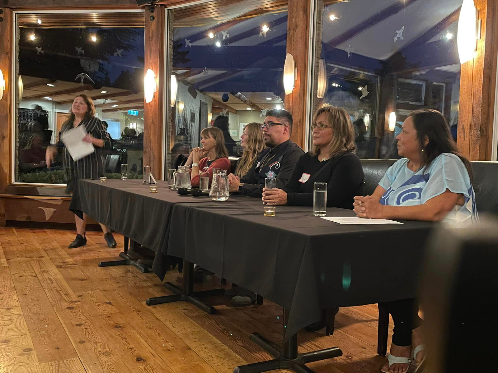 Five people sit on one side of a table covered in a black tablecloth, with water glasses and a jugs of water in
    front of them, while another person standing at the end of the table is holding a piece of paper and speaking.
    Behind them are large, wooden-framed windows.