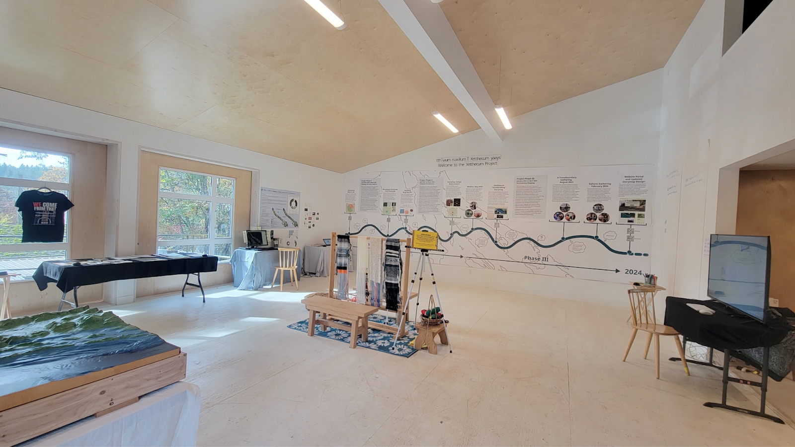 A gallery space with windows on one side and with several tables set up around the room. One table supports a
    3D map and another has a large computer screen on it, with a keyboard and chair in front. Mounted on the far wall is
    a large timeline banner taking up the entire width of the wall. A Coast Salish loom is set up in the middle of the
    room with a bench in front.