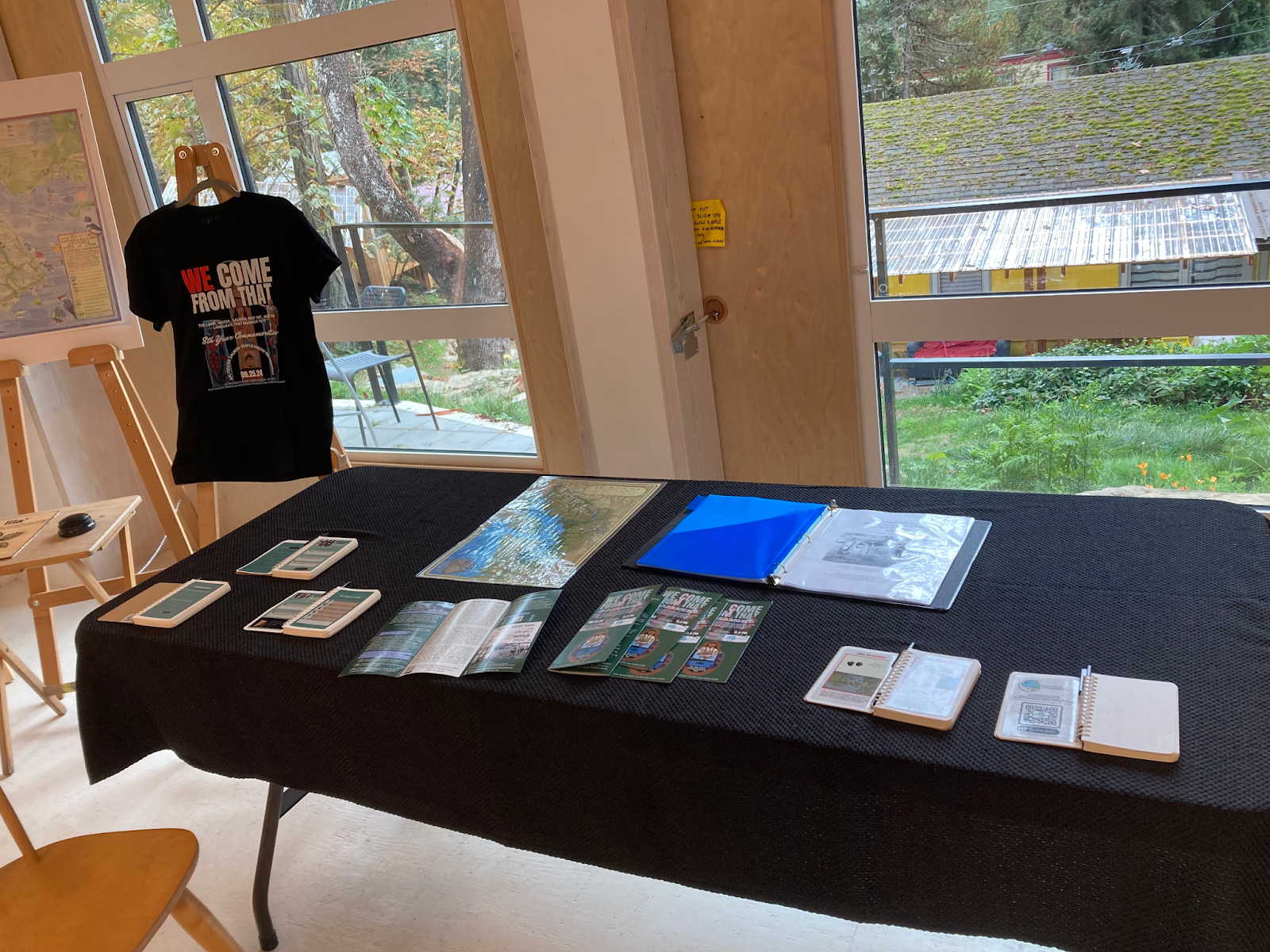 A map, binder, pamphlets and various small booklets lie on a table, beside which a T-shirt is on display
    printed with the words “WE COME FROM THAT”.